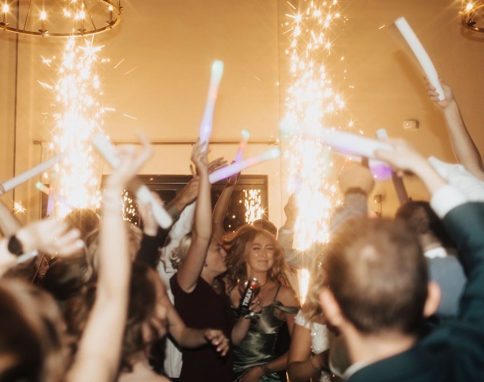 Guests celebrating with glow sticks beside cold spark fountains in a warmly lit venue.